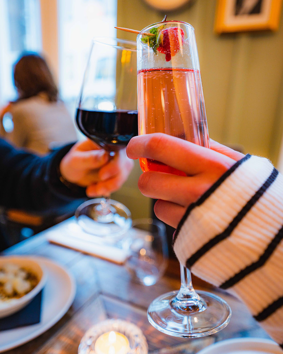 Image of a wine glass filled with red wine, cheersing with a hand held champagne flute with a sparkingling rose.